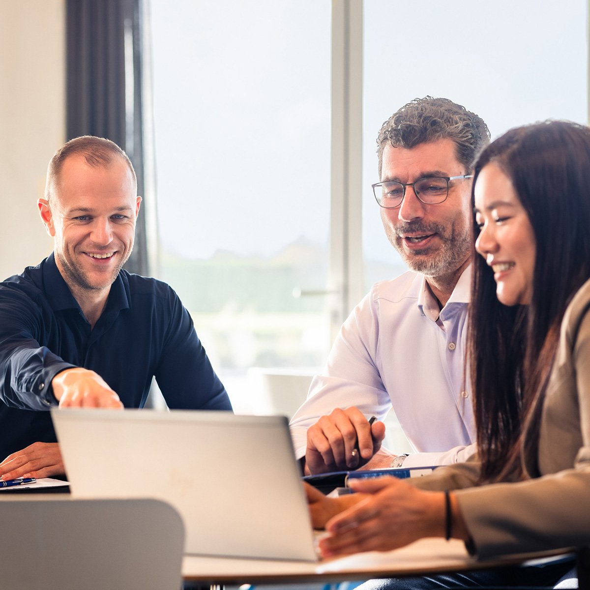 Three individuals appear to be collaborating on a laptop in a brightly lit room with a visible window in the background.