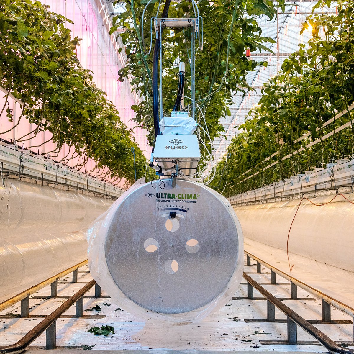 A large, circular structure with multiple circular openings on its surface, mounted on a stand with a boom and a hanging basket at the top. There are greenhouse equipment.