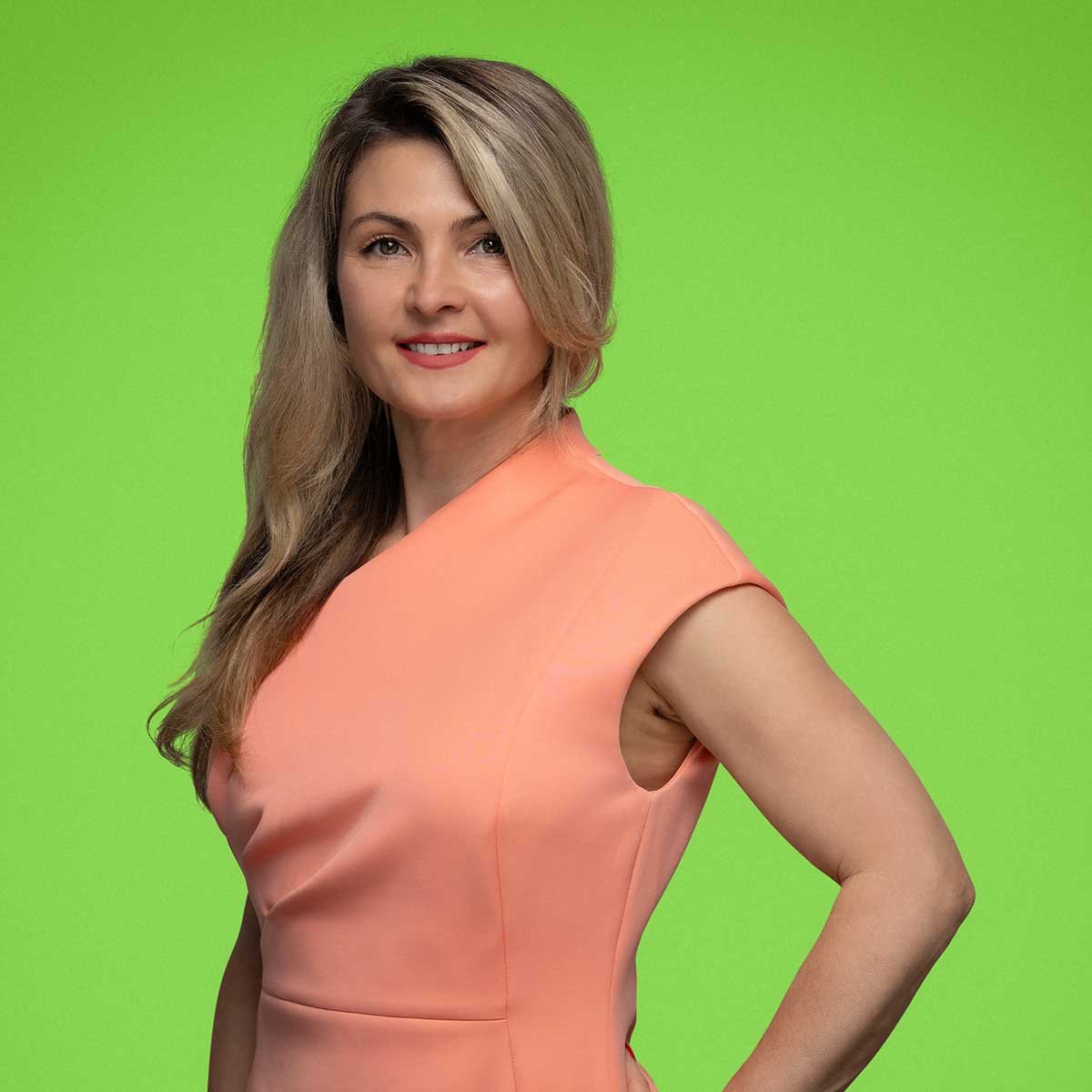 The image shows a person standing against a green background, wearing a sleeveless, light pink dress with a V-neckline and a fitted bodice, revealing the person's shoulders and arms.