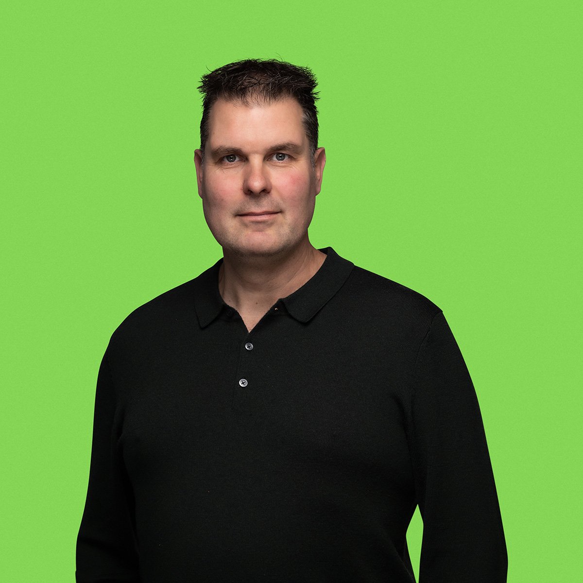 A person standing against a green background, wearing a black shirt with three buttons and long sleeves. The person's face is not visible in the image.