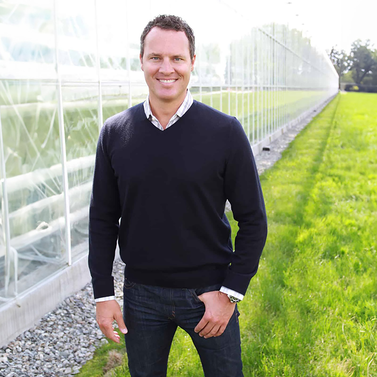 A man is standing outdoors, wearing a black sweater over a collared shirt, white shirt cuffs, jeans, and a watch on his left wrist. He has his hands in his pockets and is smiling.