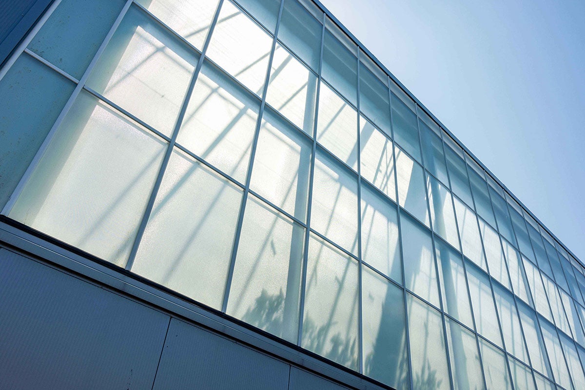 Een close-up van een modern gebouw met een gevel die bestaat uit talrijke grote, vierkante glaspanelen die in een rasterpatroon zijn gerangschikt. Het glas weerspiegelt de lucht en een deel van de structuur van het gebouw, waardoor een dynamische en eigentijdse uitstraling ontstaat. De lucht op de achtergrond is helder en blauw, wat doet vermoeden dat het een zonnige dag is.