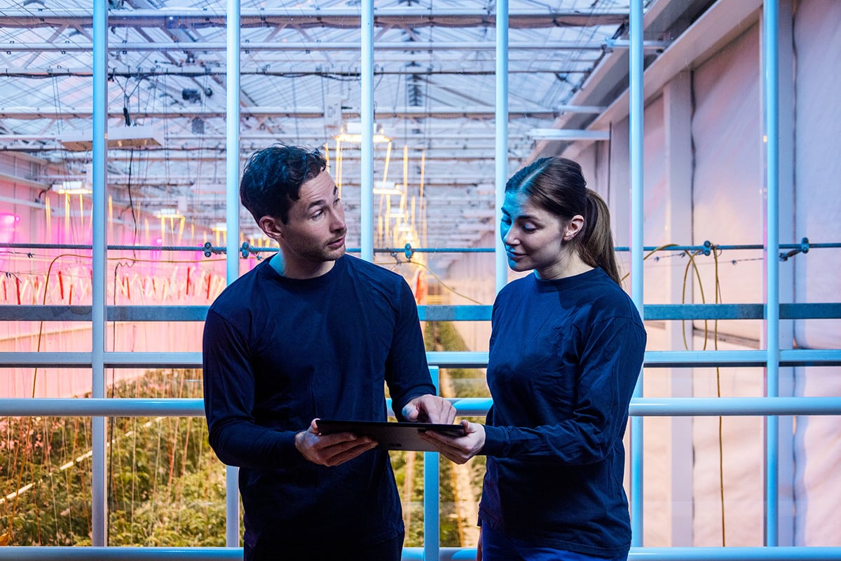 Twee personen staan op een balkon met een reling, tegenover elkaar en houden een tablet vast. De omgeving suggereert een indoor omgeving met een complex netwerk van kabels boven het hoofd en planten zichtbaar op de achtergrond.