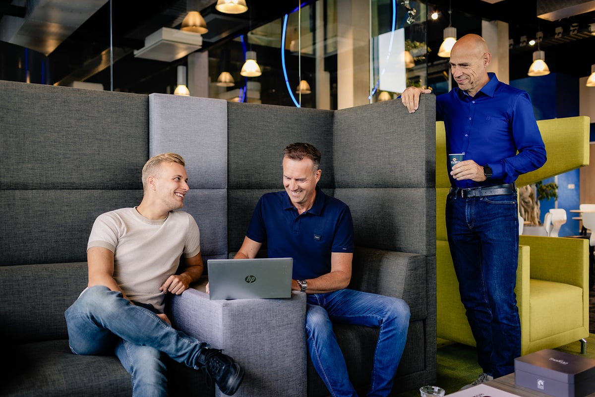Drie personen zitten op een grijze bank in een modern interieur. Twee zijn geconcentreerd op een laptop, terwijl de derde, staand, een drankje vasthoudt.