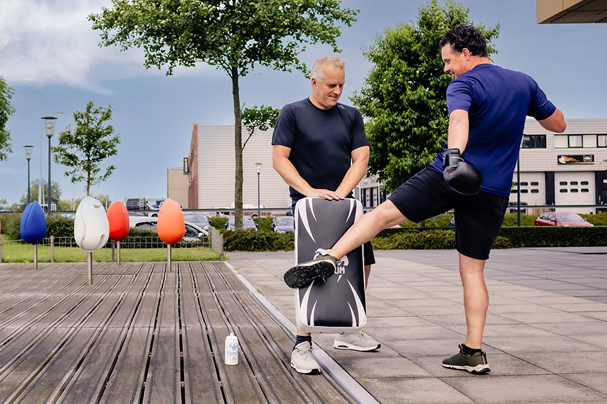 Two individuals engaged in a boxing training session on a wooden deck. The person on the right is in the midst of delivering a kick with their right leg extended, while the person on the left is crouched down. Both individuals are wearing boxing gloves and are focused on the training exercise.