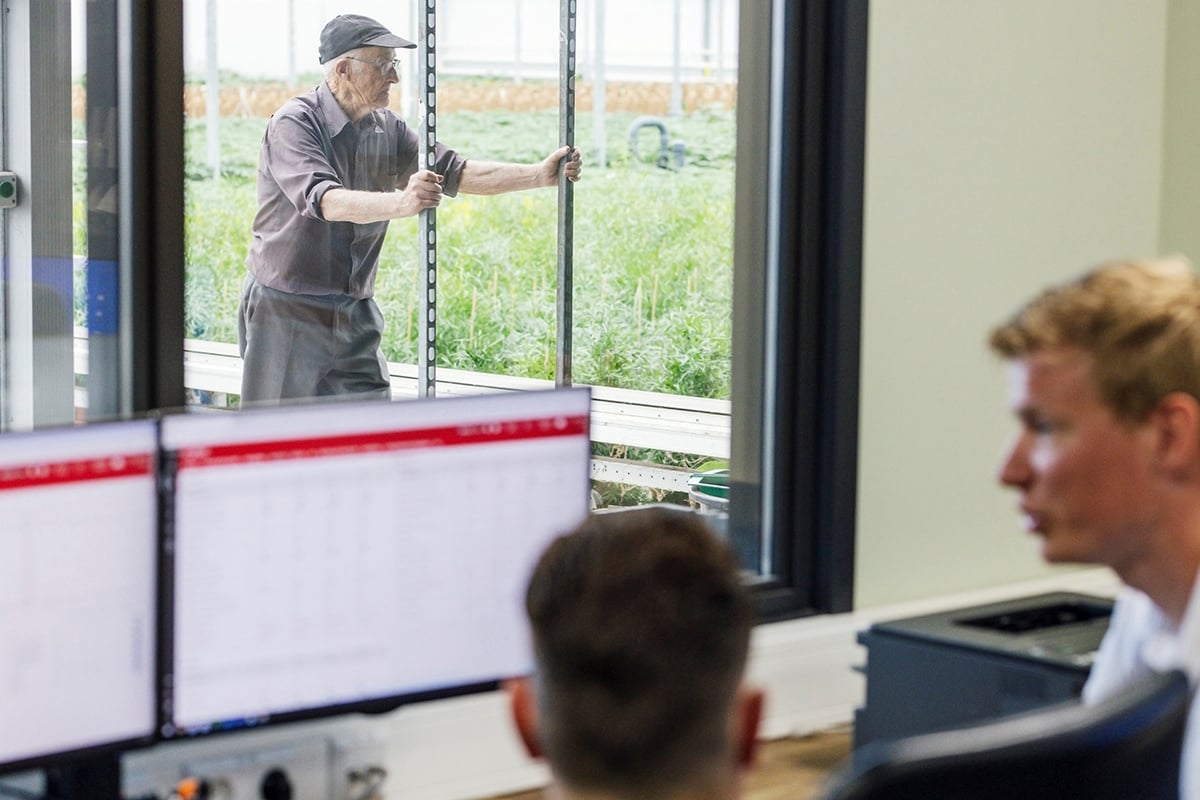 An indoor setting where three individuals are present. The person in the foreground is sitting at a workstation with multiple computer monitors. The person in the background is standing by a window with a view of greenery outside.