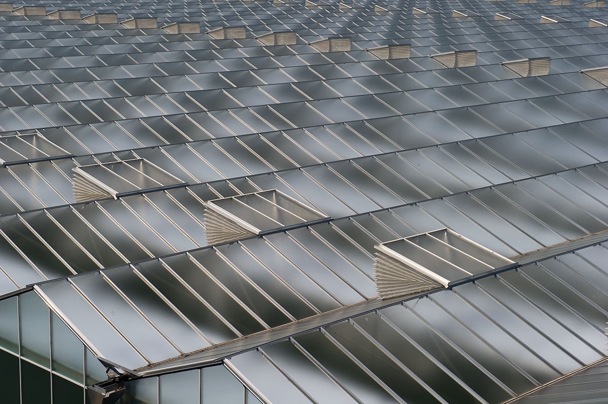 De afbeelding toont een dak van een moderne kas. Het dak heeft een reflecterend oppervlak, waardoor een glinsterend effect ontstaat. Tussen sommige glazen ramen zitten openingen of ventilatieopeningen. De hoek van de afbeelding is van onderen, omhoog kijkend naar de bovenkant van het gebouw.