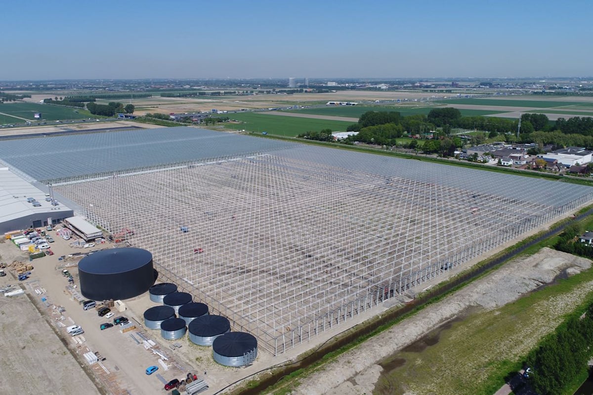 An aerial view of a large greenhouse. The facility is surrounded by fields and some buildings in the background. There are also storage tanks and construction materials visible near the facility.