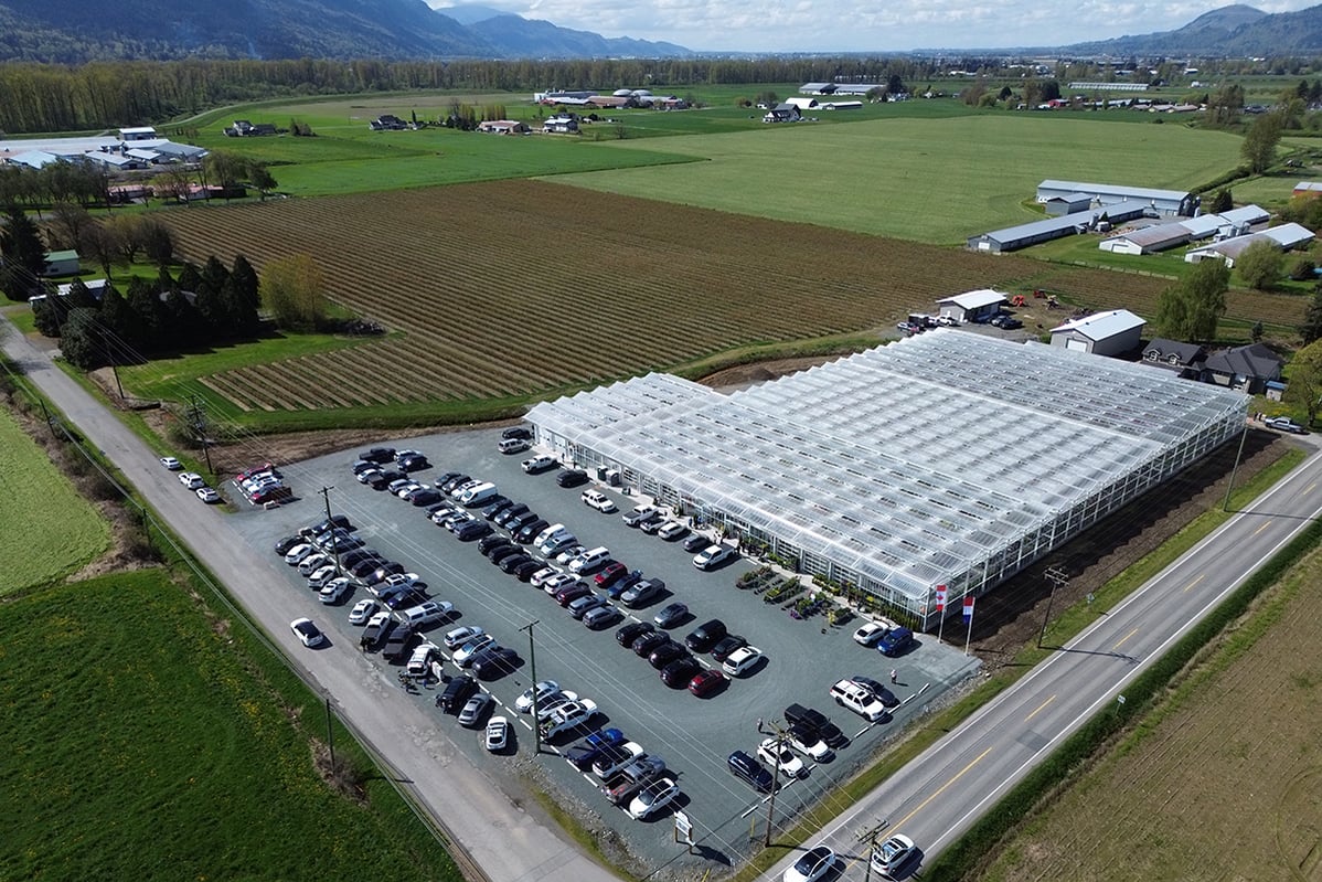 An aerial view of a large industrial facility with a greenhouse structure surrounded by a parking lot filled with cars. In the background, there is an agricultural field with rows of crops and a few buildings.
