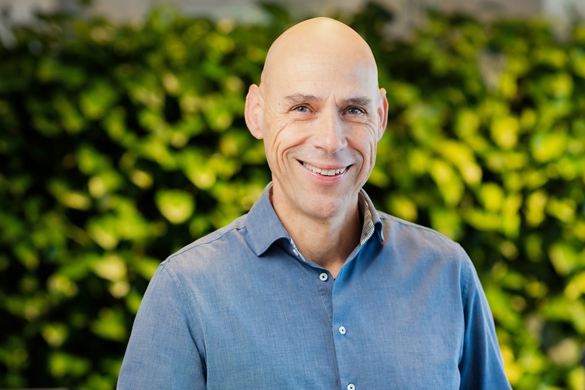 A bald man with a shiny smile, wearing a blue shirt, poses in front of a background of green leaves.
