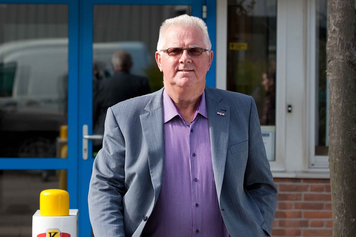 A person is standing outdoors wearing a grey blazer over a purple shirt, with a tree on the left and a blue door in the background.