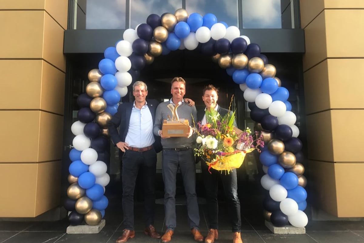 Three individuals standing in front of an entrance decorated with blue and white balloons and gold accents. The person in the center is holding a large bouquet of flowers and a box, possibly indicating a celebration or event.
