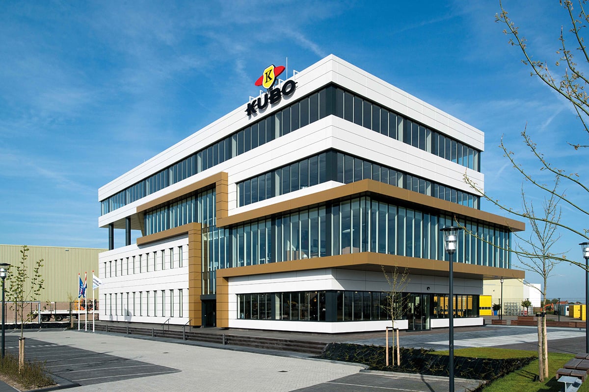 A modern multi-story building with a combination of white and dark brown facade. The top of the building features a logo with the word 'fuso'. The building has large glass windows and is situated on a paved area with a few trees and street lamps. The sky is clear and blue.