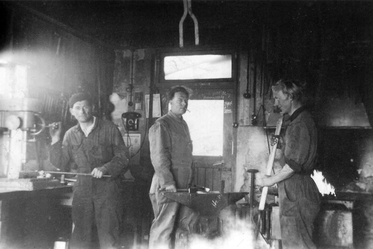 Een zwart-wit foto van vier personen in een vroeg 20ste-eeuwse werkplaats. Ze zijn gekleed in werkkleding uit die tijd, inclusief overalls en hoeden, en lijken bezig met een taak waarbij machines of apparatuur betrokken zijn.
