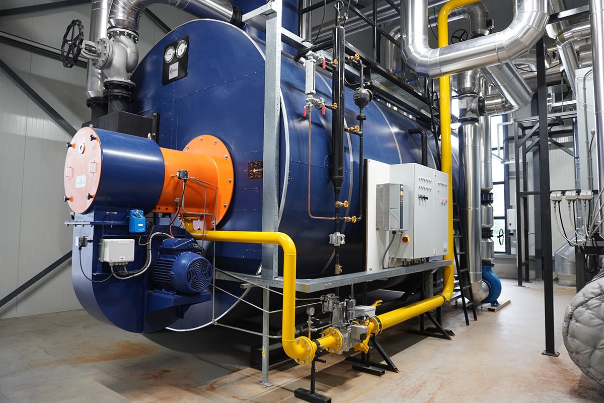 Een industriële omgeving met een grote blauwe stoomketel als middelpunt. De ketel is voorzien van een oranje veiligheidsklep bovenop en er zitten meerdere pijpen en kleppen omheen, wat doet vermoeden dat hij deel uitmaakt van een groter verwarmings- of stroomopwekkingssysteem.