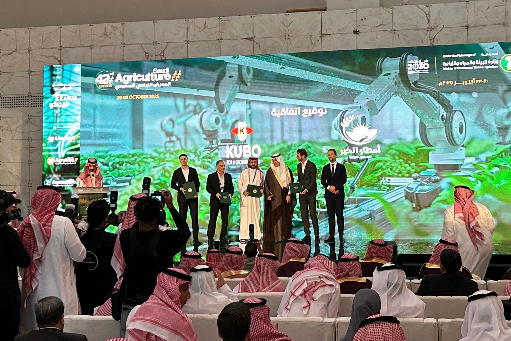 مجموعة من الأشخاص يقفون على خشبة المسرح أمام شاشة كبيرة تعرض عرضًا تقديميًّا حول موضوع الزراعة. ويجلس الجمهور في صفوف مواجهة خشبة المسرح.