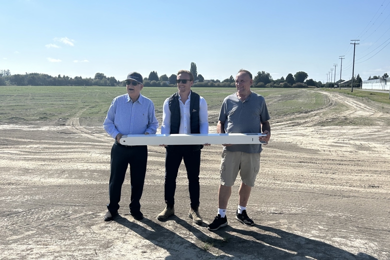 Three men are standing on an open construction site, holding a long white object together